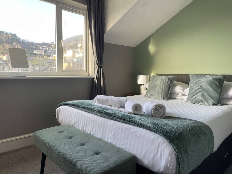 Un letto rifatto con una coperta a righe verdi e bianche, una panca rivestita di verde e vista su una zona residenziale attraverso una finestra.