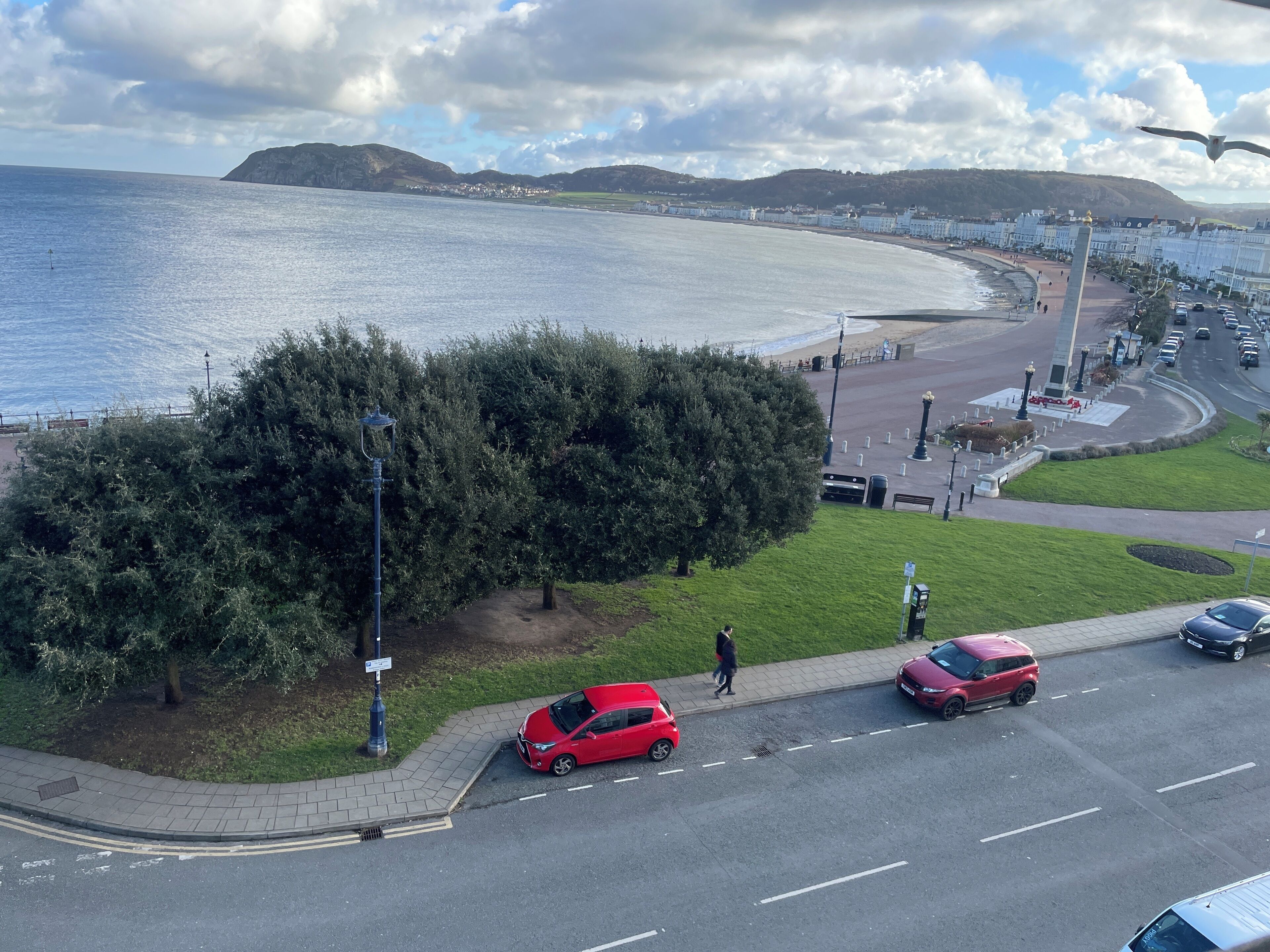 A coastal road with a red car parked on the side, a pedestrian crossing, and a view of the sea and hills in the background.