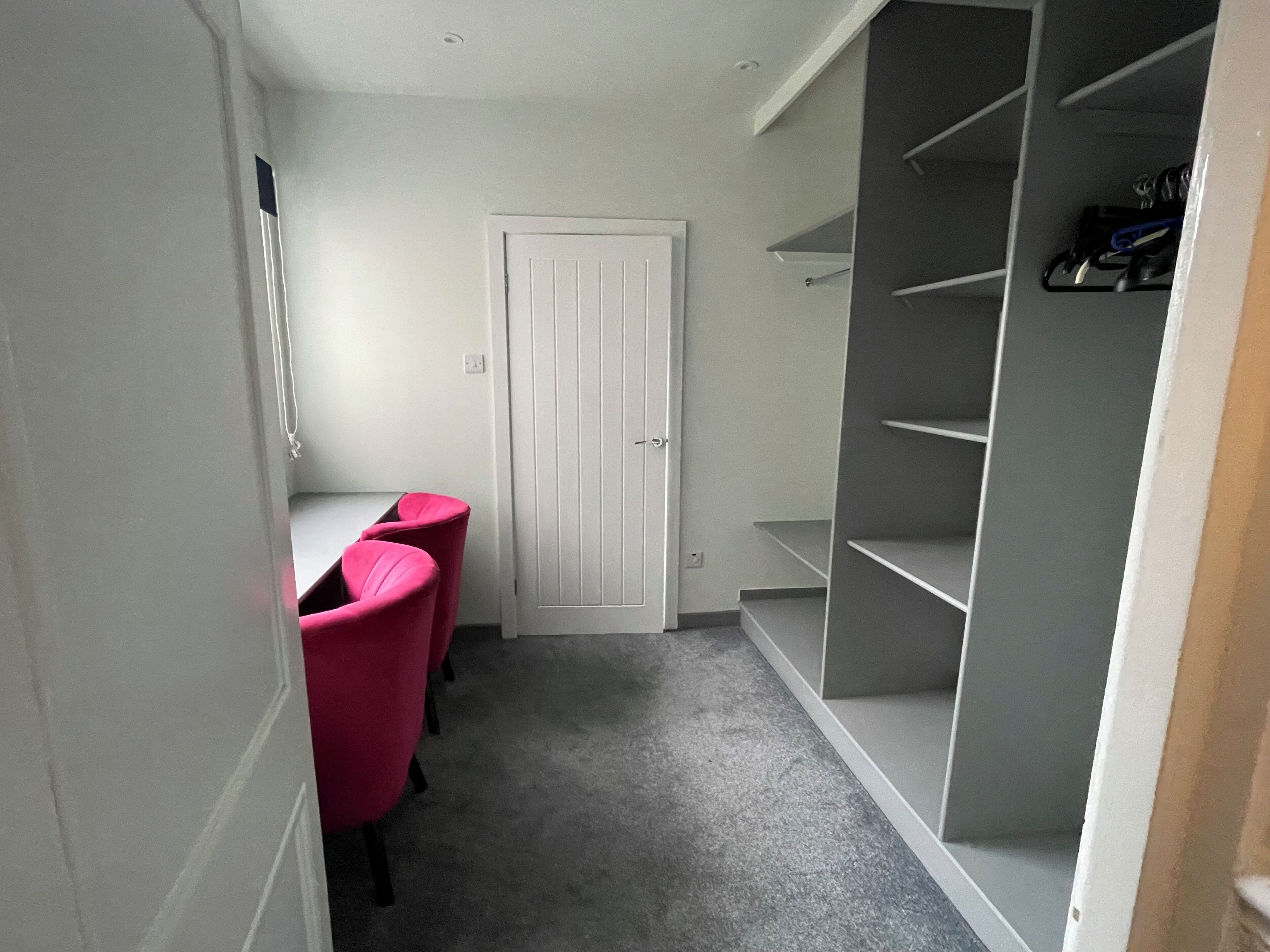 A narrow hallway with a white door, a grey carpet, and a grey shelving unit with shoes on the right.