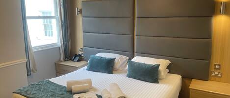 A neatly made bed with a quilted green bedspread, two pillows, and rolled towels. There is a bedside table with a phone and a lamp.