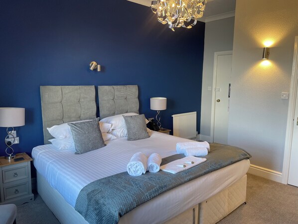 A neatly made bed with white linens and grey pillows, a bedside table with a lamp, and a wall-mounted light fixture.