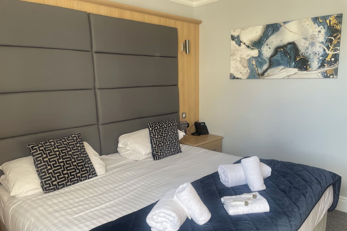 Hotellihuone, jossa on suuri sänky, puinen pääty, yöpöytä, jossa on puhelin, ja seinällä taulu.