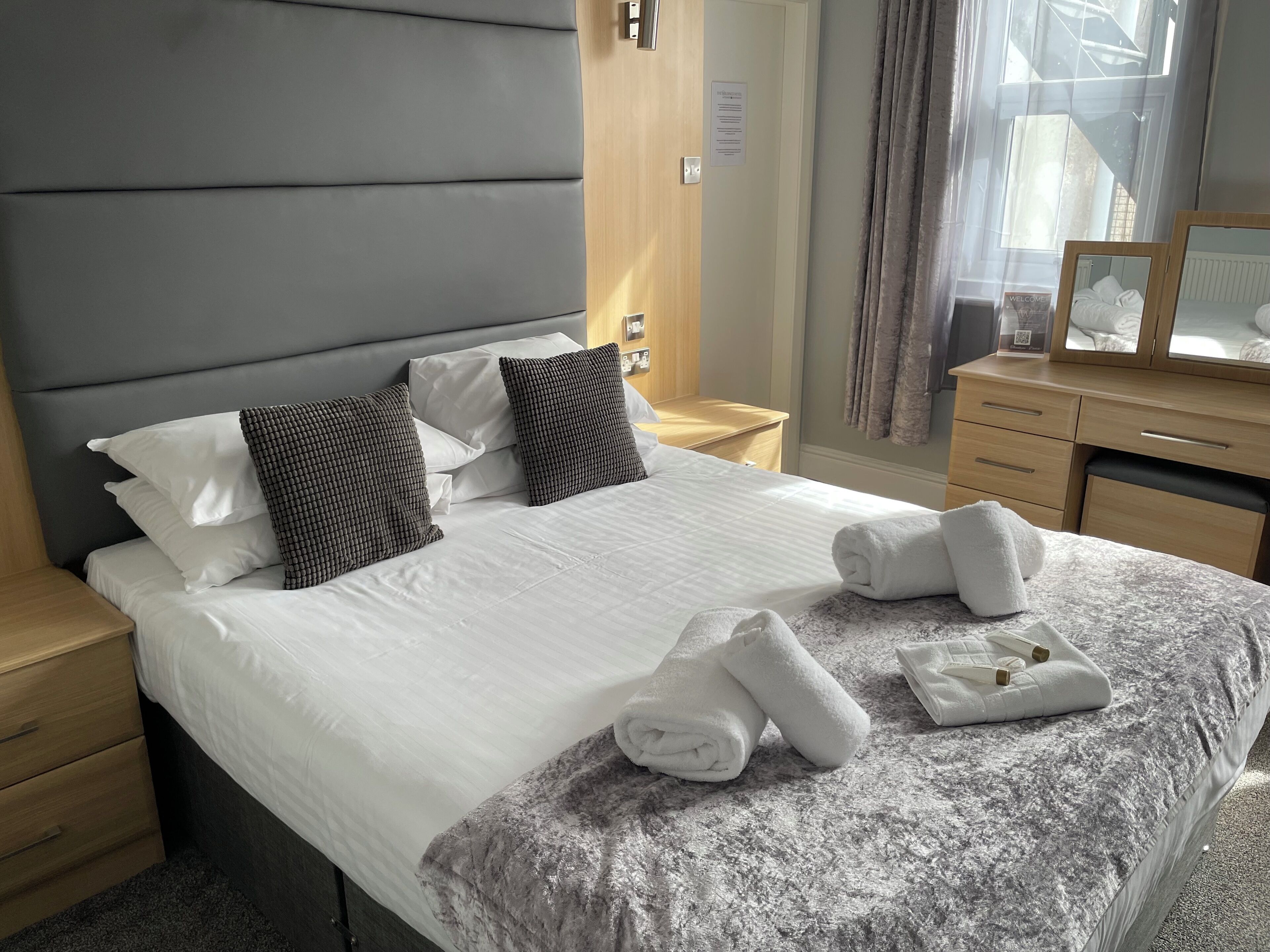 Una cama bien hecha con sábanas blancas y un edredón gris con textura, una mesita de noche de madera con una lámpara y un aparador de madera con espejo.