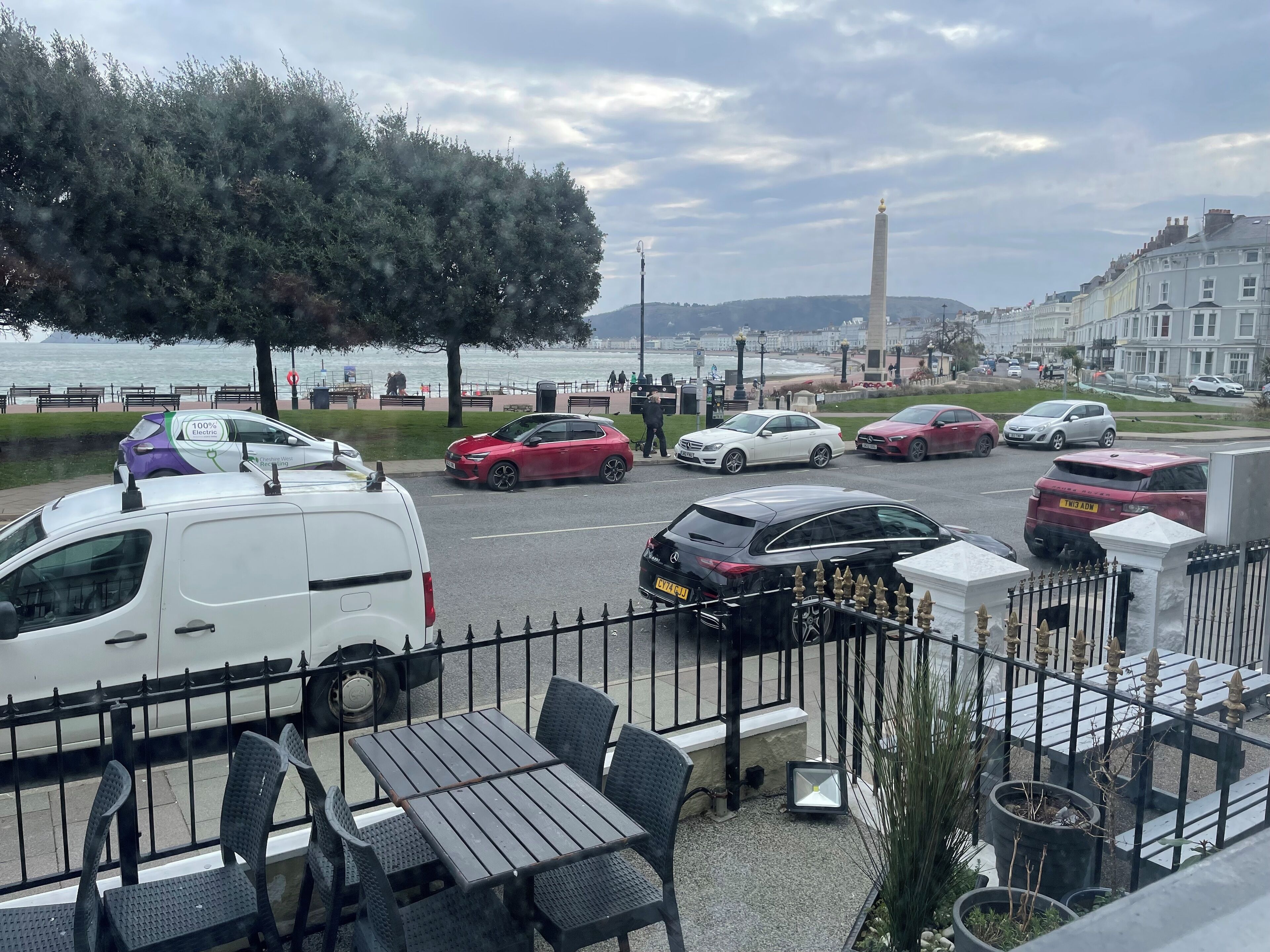 A coastal view with parked cars, a white van, and a table with chairs.