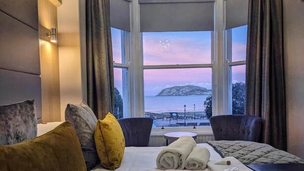 A hotel room with a large bay window, a bed with rolled towels, a small round table, and two armchairs. - The Wildings (Llandudno)