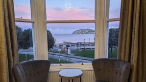 A room with two armchairs and a small table, a large window with a view of a pier and beach, and curtains on either side. - The Wildings (Llandudno)