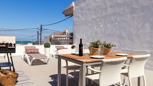Eine Dachterrasse mit Holztisch, weißen Korbstühlen und Blick aufs Meer.