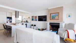 Ein modernes Wohnzimmer mit Kamin, einem Essbereich und einer dahinterliegenden Küche. Ein großes Fernsehgerät ist an der Wand befestigt.