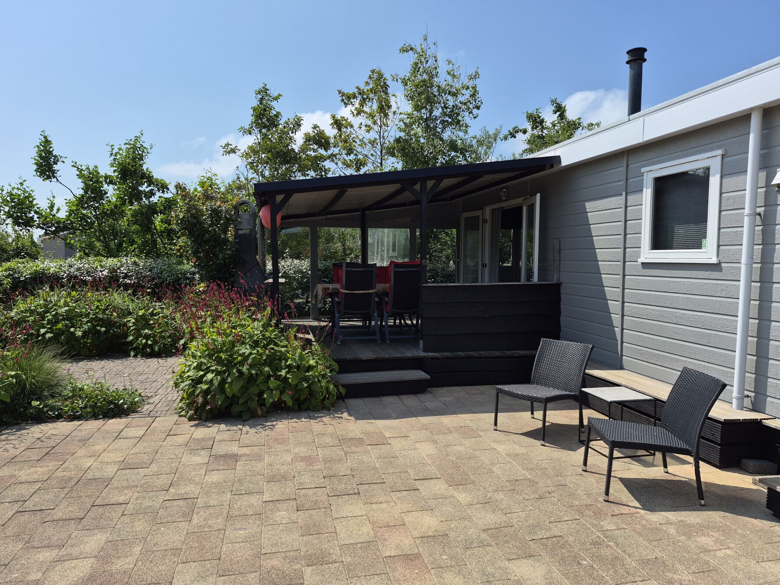 Une terrasse extérieure couverte, dotée d’une table et de chaises, et entourée d’un jardin où poussent différentes plantes, ainsi que d’un chemin en briques.