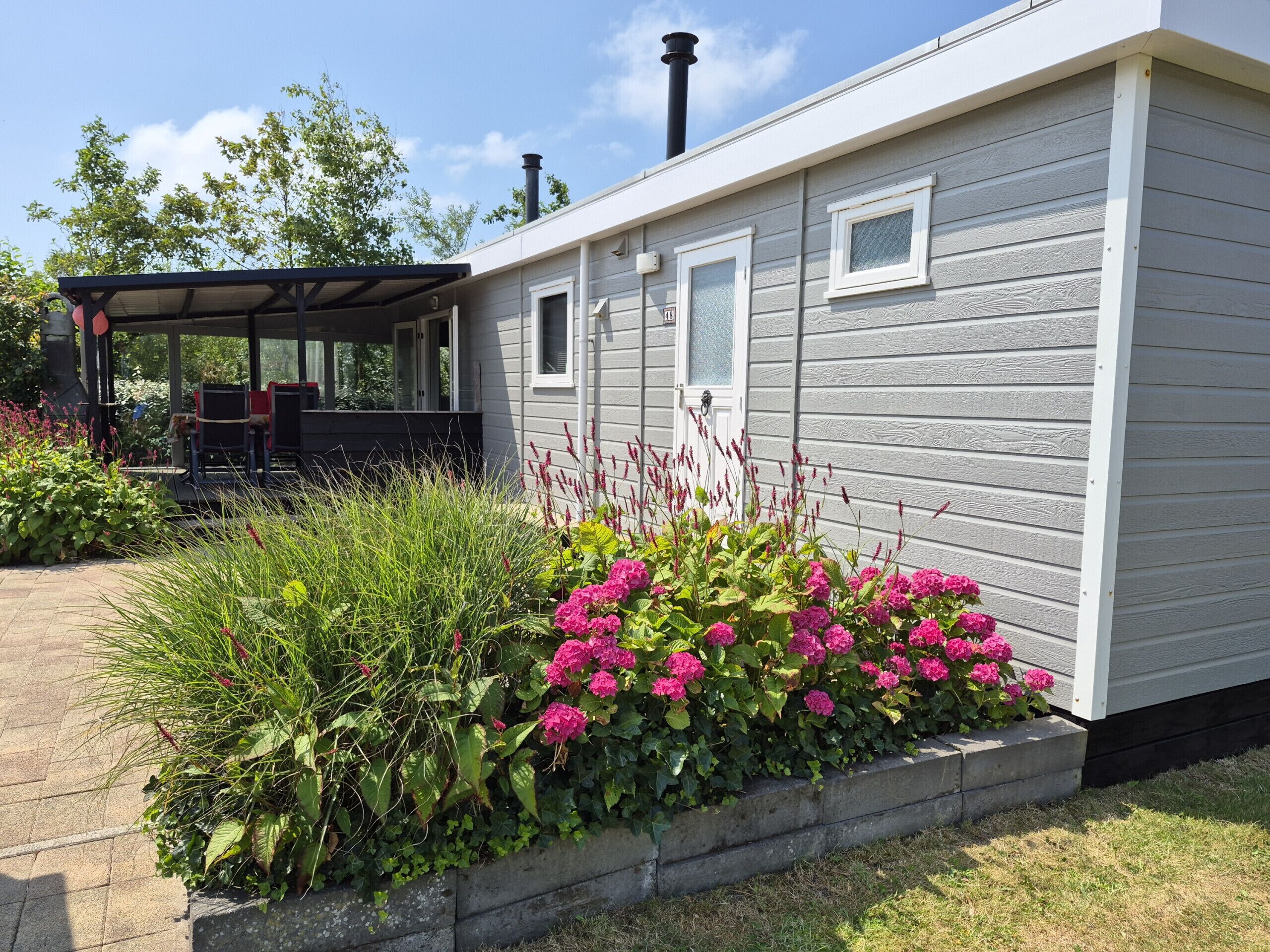 Une maison de plain-pied avec un porche couvert, une cheminée et un jardin agrémenté de fleurs roses.