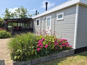 A single-story house with a covered porch, a chimney, and a garden with pink flowers. - CS 48 - Callassande Vacation Park (Callantsoog)