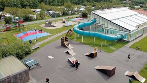 A playground with various play structures, a large building with a green roof, and a swimming pool. - CS 48 - Callassande Vacation Park (Callantsoog)