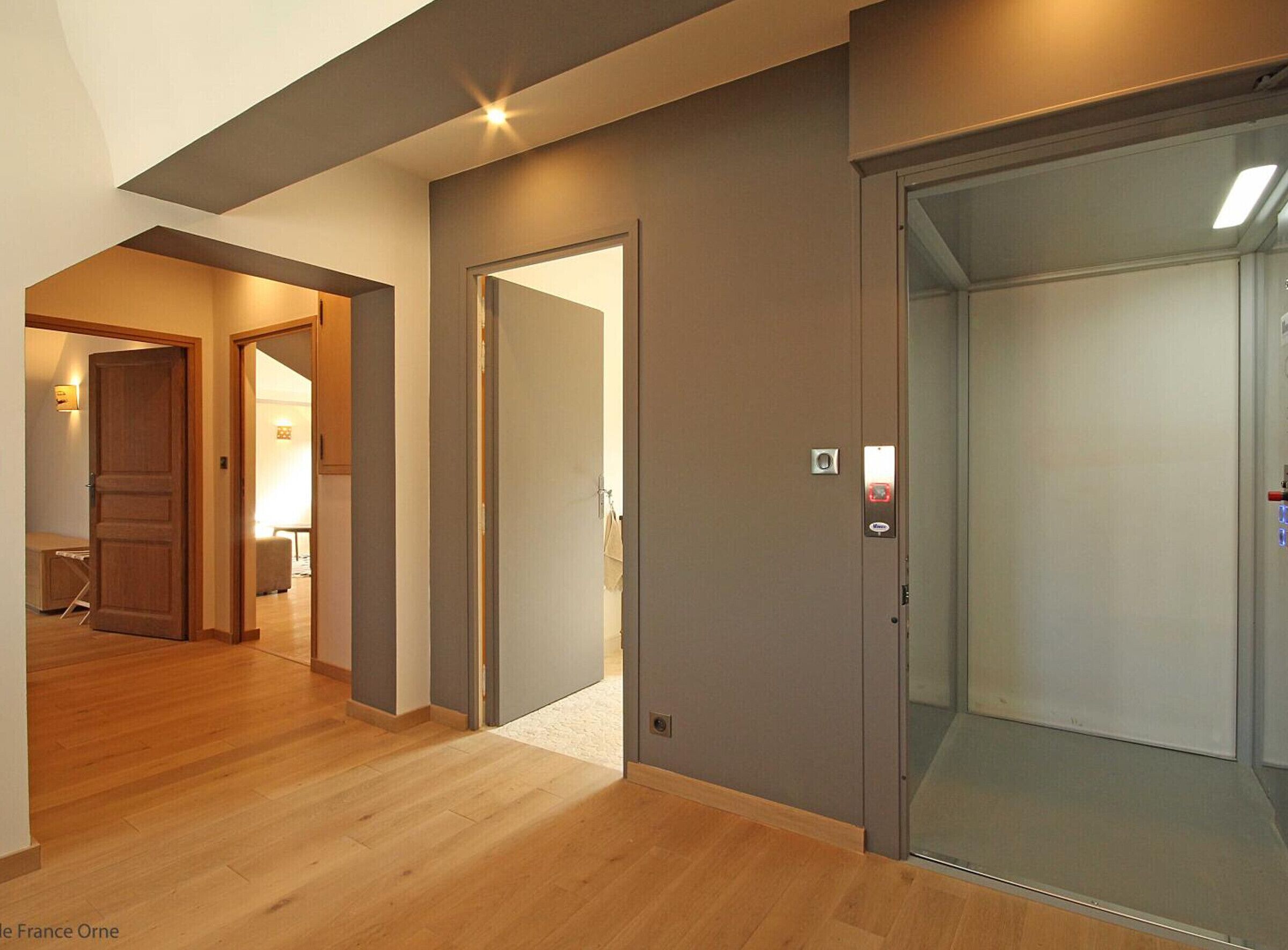 Un intérieur moderne avec un plancher en bois, un couloir menant à une pièce avec un lit, et un ascenseur.