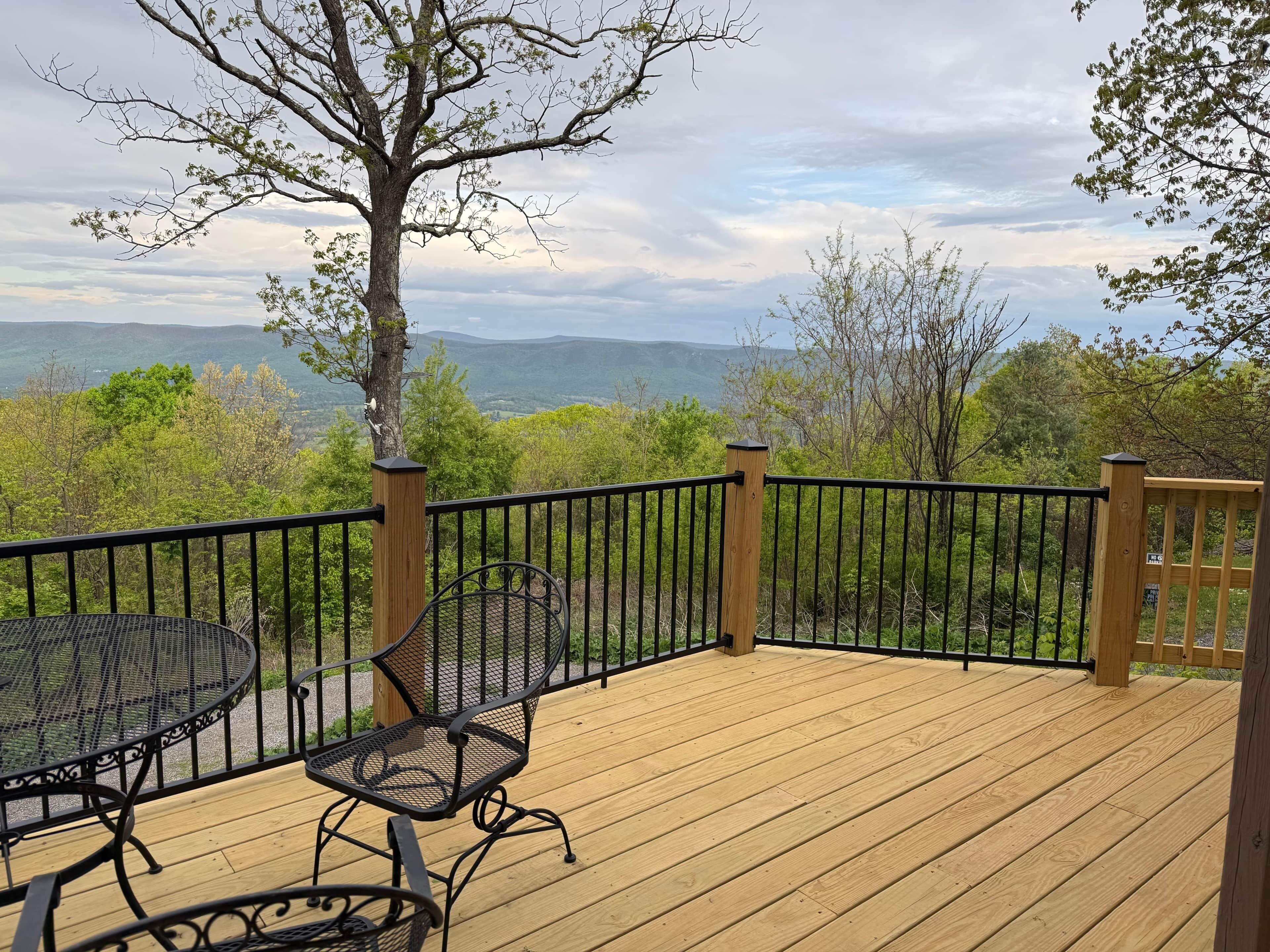 Une terrasse en bois, agrémentée d’une table et de chaises, entourée d’une rambarde en métal noir, et offrant une vue sur un paysage montagneux.