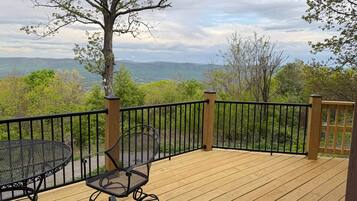 Une terrasse en bois, agrémentée d’une table et de chaises, entourée d’une rambarde en métal noir, et offrant une vue sur un paysage montagneux.
