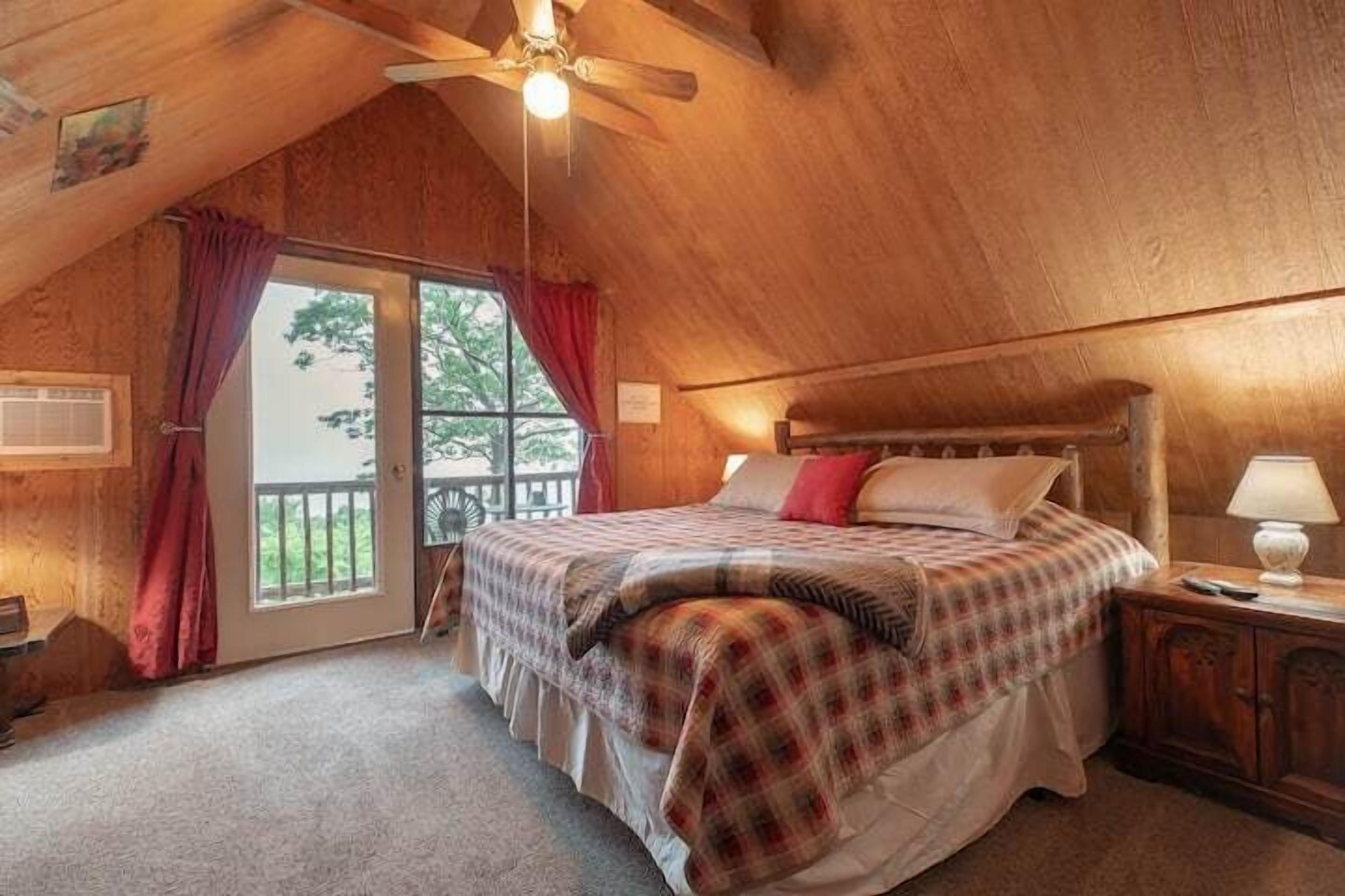 Een gezellige slaapkamer met een houten plafond, een bed met een geruit sprei, een nachtkastje met een lamp en een glazen schuifdeur naar een buitenruimte.