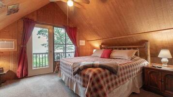 Een gezellige slaapkamer met een houten plafond, een bed met een geruit sprei, een nachtkastje met een lamp en een glazen schuifdeur naar een buitenruimte.