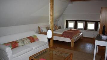 Um quarto acolhedor com um sofá branco, uma mesa de centro de madeira, uma cama com manta vermelha e uma pequena mesa com um rádio.