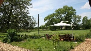 Un área de estar al aire libre con una mesa y sillas bajo un paraguas blanco, rodeada de un campo cubierto de césped y árboles.