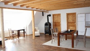 Una habitación amplia con vigas de madera, chimenea, comedor con sillas y mesa, y zona de cocina con armarios y un refrigerador.