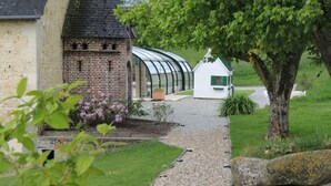 A pathway leading to a brick building with a glass greenhouse, surrounded by greenery and a white house. - Spacious House with Indoor Pool and Wifi in Laleu (Laleu)