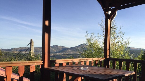 Un pontile in legno con panche e un tavolo, affacciato su un paesaggio montuoso con un ponte sullo sfondo.