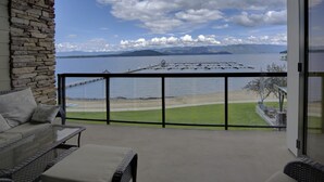 A balcony with a stone wall, a glass door, and a view of a lake with mountains in the background. - A Scenic Canvas, Lakefront - Seasons at Sandpoint (Sandpoint)