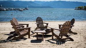 A beach with three wooden Adirondack chairs and a fire pit, overlooking a lake with boats and mountains in the background. - A Scenic Canvas, Lakefront - Seasons at Sandpoint (Sandpoint)
