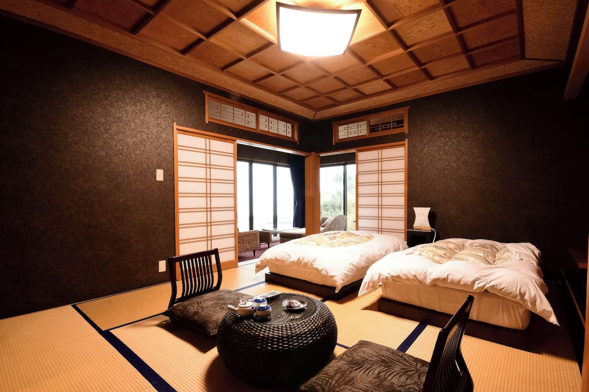 A traditional Japanese-style bedroom with tatami flooring, two beds, a futon, a low table with a tea set, and sliding doors.