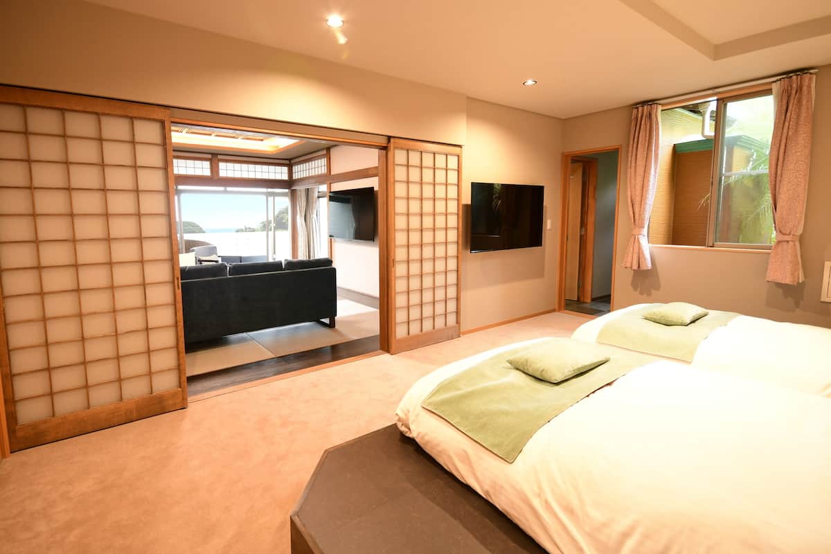 A hotel room with a sliding shoji screen, a bed with green pillows, a TV mounted on the wall, and a view of the outdoors.