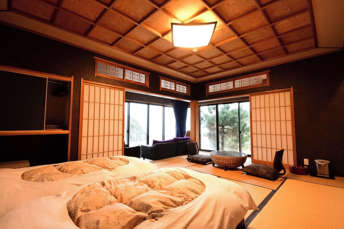 A traditional Japanese-style room with tatami flooring, a large bed with pillows, a futon, a low table, and sliding doors.