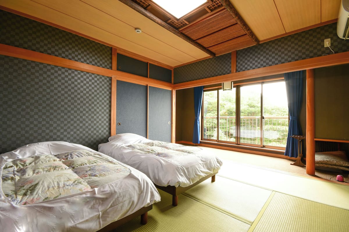 A traditional Japanese room with tatami flooring, two beds with floral bedding, a large window with blue curtains, and a view of greenery.