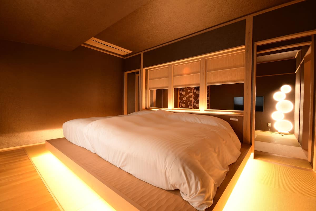 A neatly made bed in a room with a large window, a bedside table, and a mirror with lights.