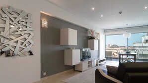 A modern living room with a flat-screen TV, a decorative wall piece, and a view of the city through large windows.
