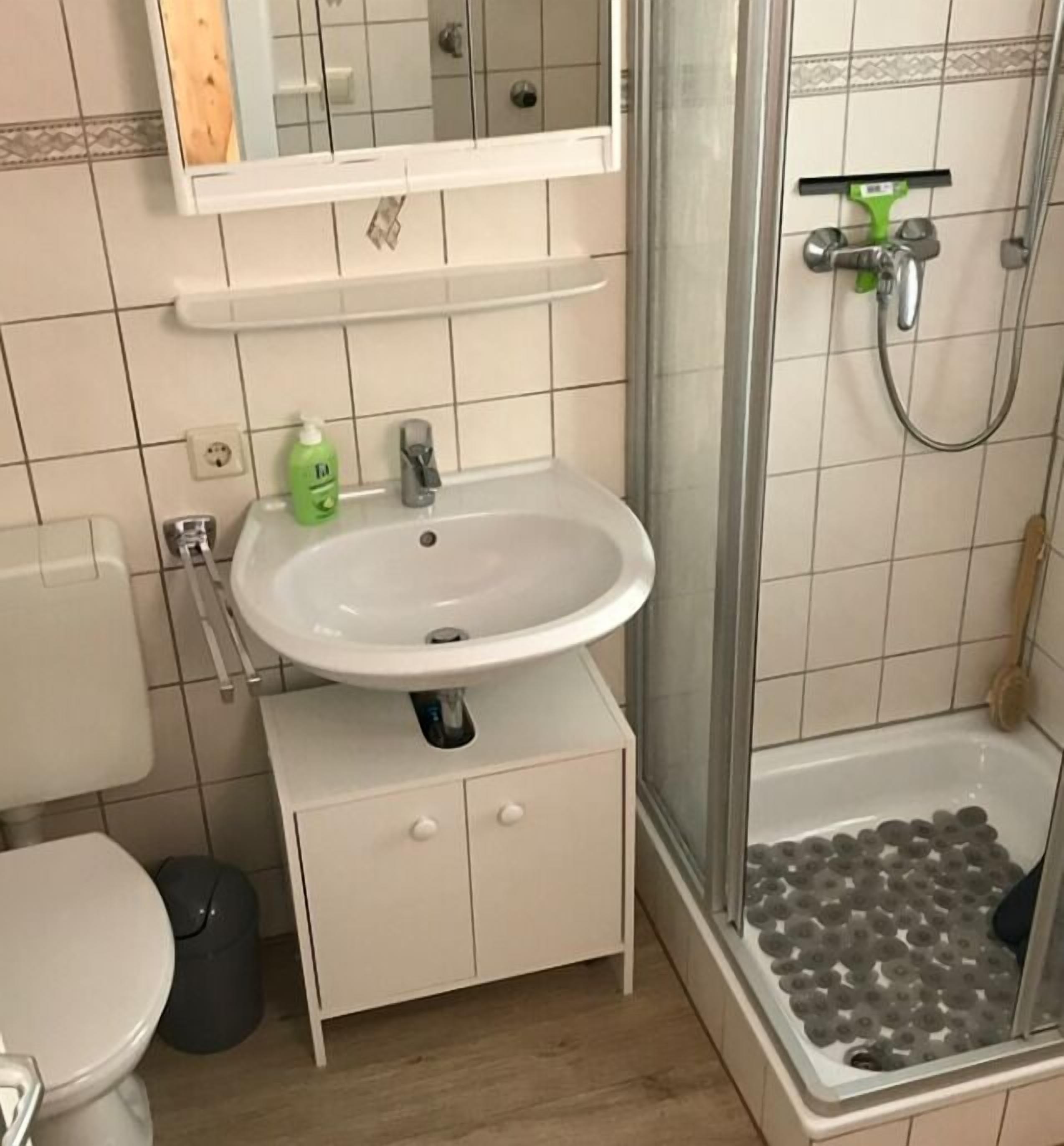 Un bagno con un lavandino bianco, uno specchio sopra, una doccia con porta in vetro e un pavimento piastrellato.
