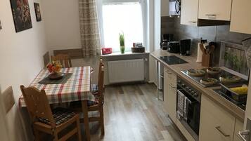 Une cuisine de taille modeste avec un coin repas, comprenant une nappe à carreaux, des chaises en bois, un radiateur, une cuisinière et une fenêtre avec des rideaux.