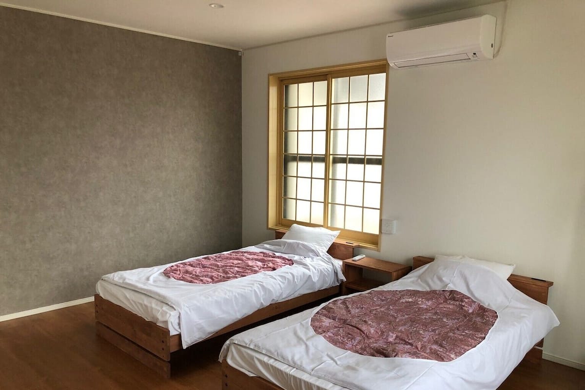 Une chambre avec deux lits en bois, une fenêtre avec des stores et un appareil de climatisation.