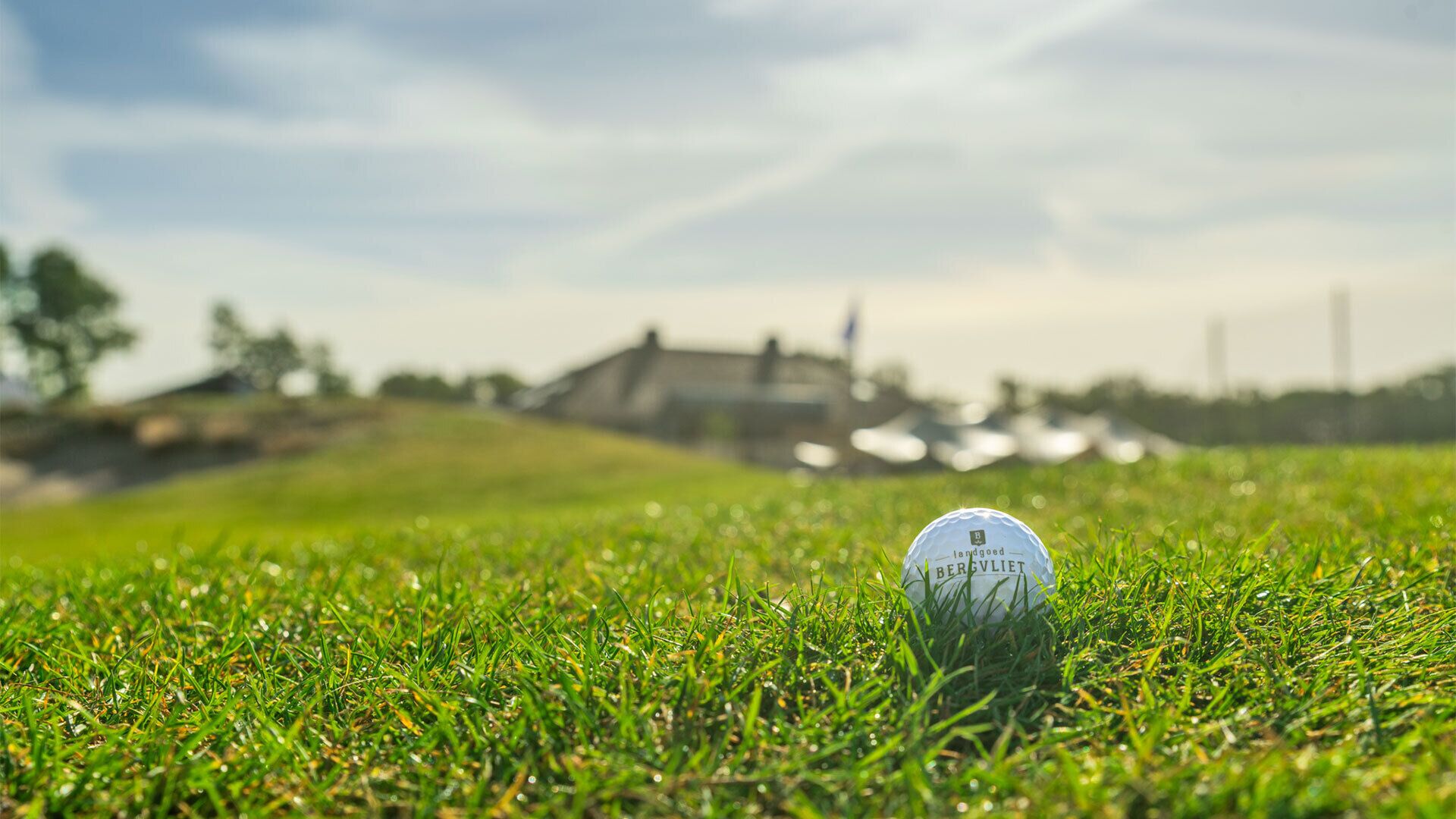 Une balle de golf sur un terrain d’herbe bien entretenu, avec un bâtiment et une étendue d’eau visibles au loin, en arrière-plan.