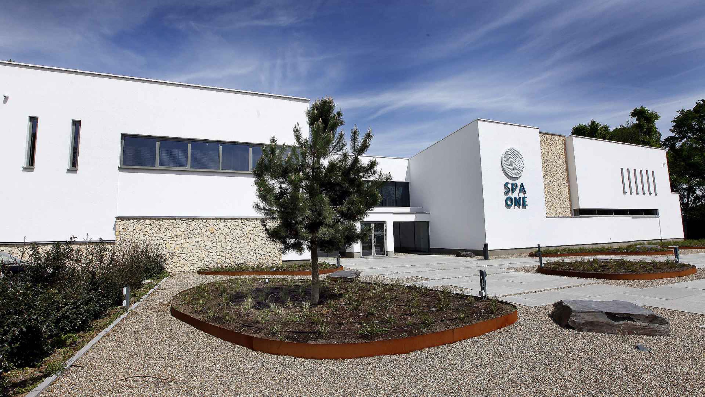A modern building with a spa logo, a stone wall, and a landscaped area with a tree.