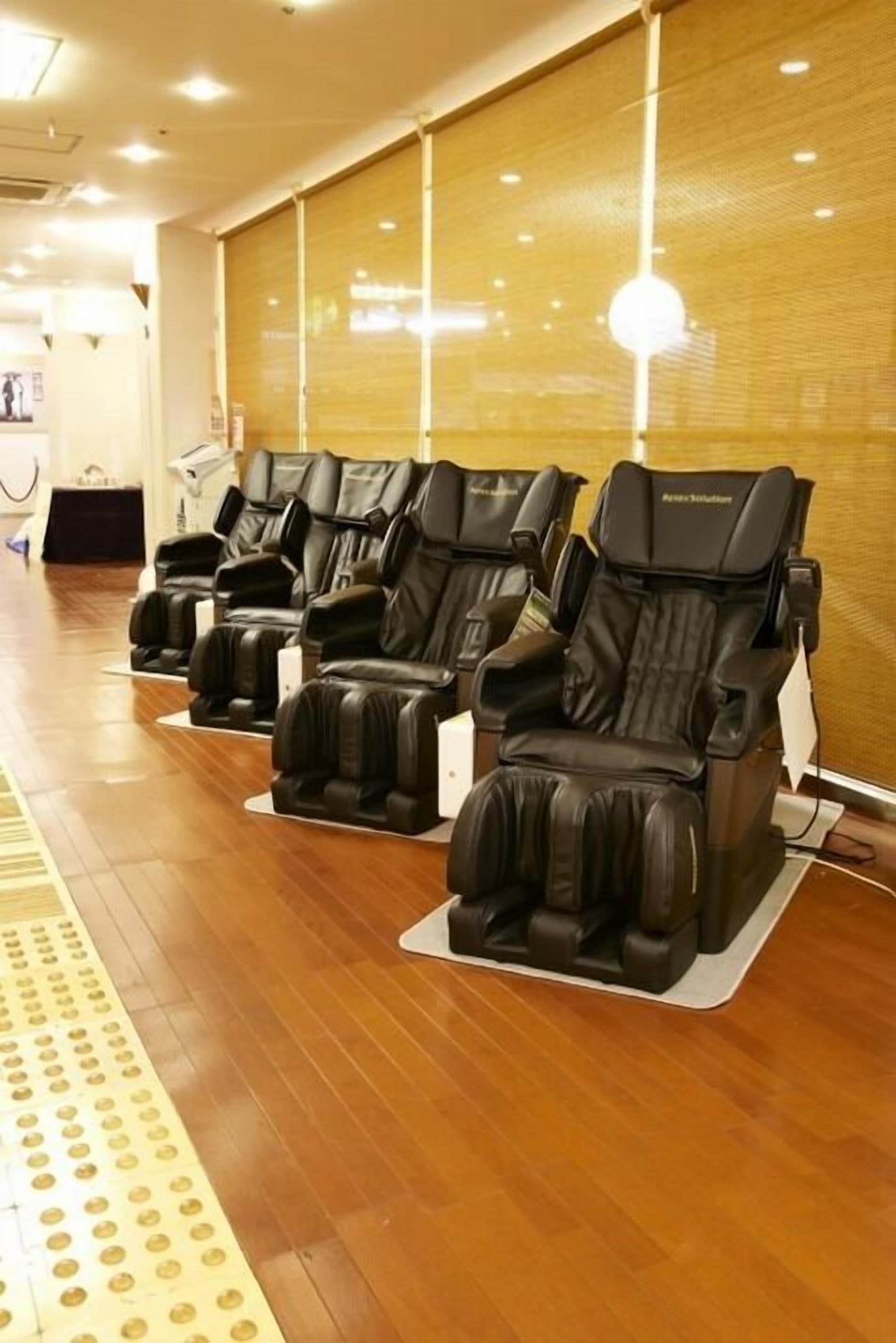 A row of massage chairs in a room with wooden flooring and a large window with blinds.