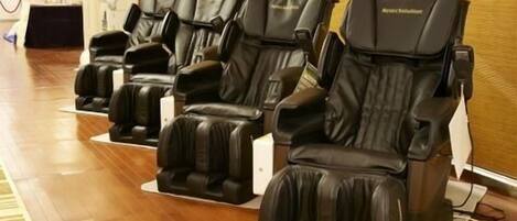 A row of massage chairs in a room with wooden flooring and a large window with blinds.