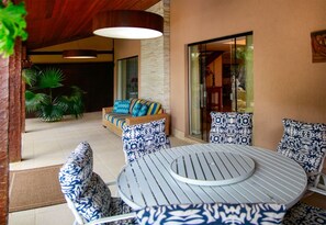A patio area with a round table and chairs, a sofa with patterned cushions, and a large window. - Villa Puerto Nuevo (Búzios)