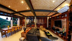 A spacious living area with a large sofa, dining table, and bar seating. There is a TV mounted on the wall and a fireplace. - Villa Puerto Nuevo (Búzios)