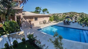 A swimming pool with lounge chairs and a patio area, surrounded by greenery and a view of a hillside town. - Villa Puerto Nuevo (Búzios)