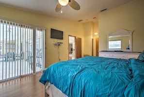 Quarto com cama grande, televisão na parede, espelho sobre a cômoda e porta de vidro corrediça para área externa.