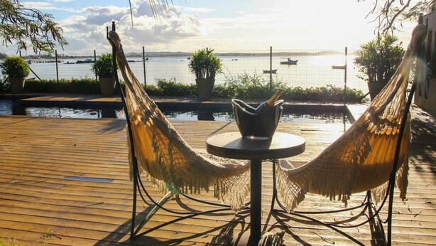 A hammock chair and table set on a wooden deck with a view of the sea and boats. -  Villa Manguinhos (Búzios)