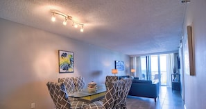 A living room with a sofa, two armchairs, a coffee table, and a dining table. There are two wall-mounted lamps, a painting, and a window with curtains.