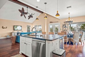 A spacious kitchen with a central island, pendant lighting, and a dining area with wicker chairs. - Lakefront Beach Home in Cinnamon Shore w/ Vaulted Ceilings & Amenity Access (Port Aransas)