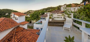 A rooftop terrace with a white chair, a small pool, and a view of a hillside with houses.