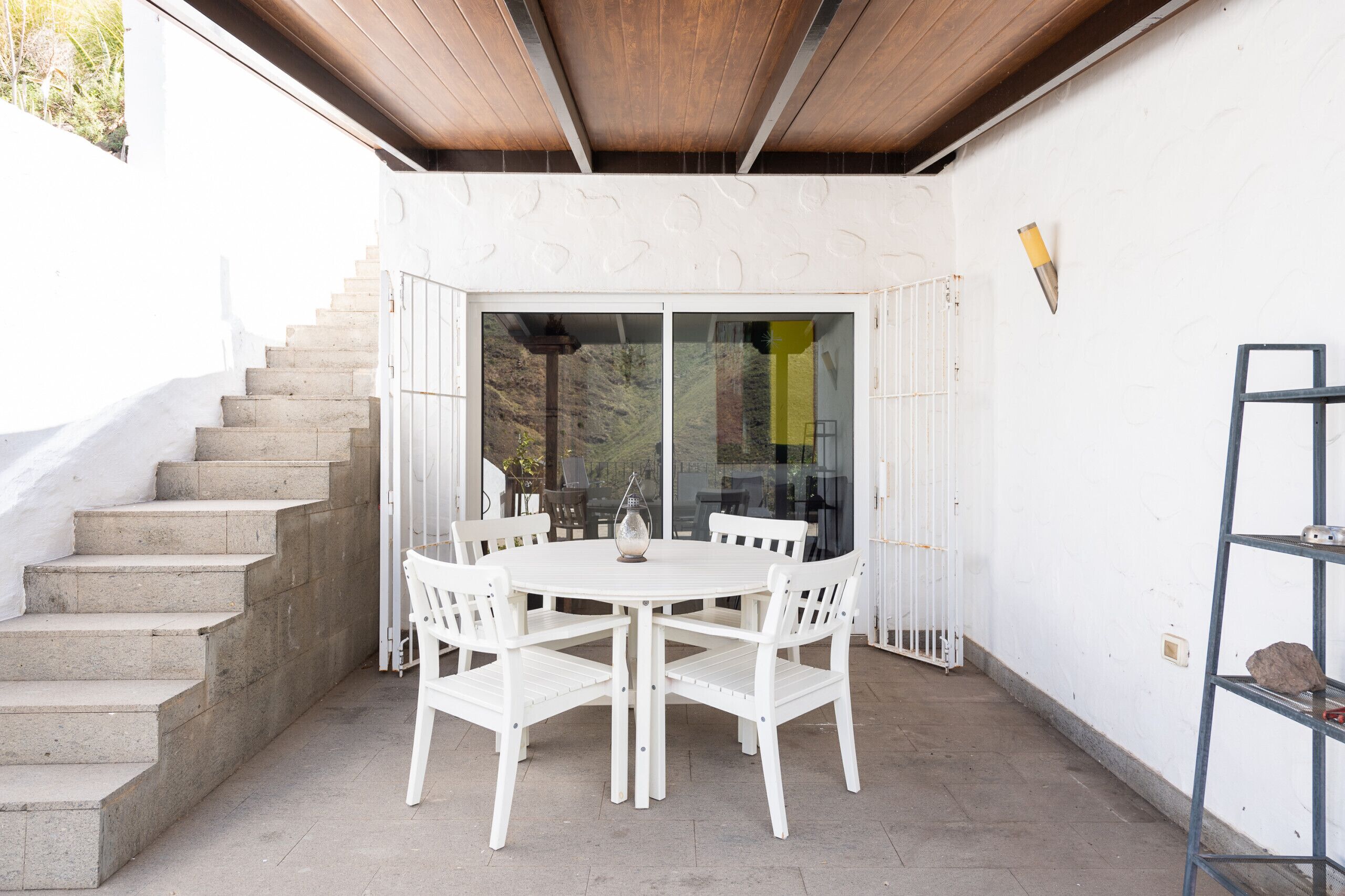 Un patio exterior con mesa y sillas redondas, una escalera y un ventanal de vidrio que da a un jardín.